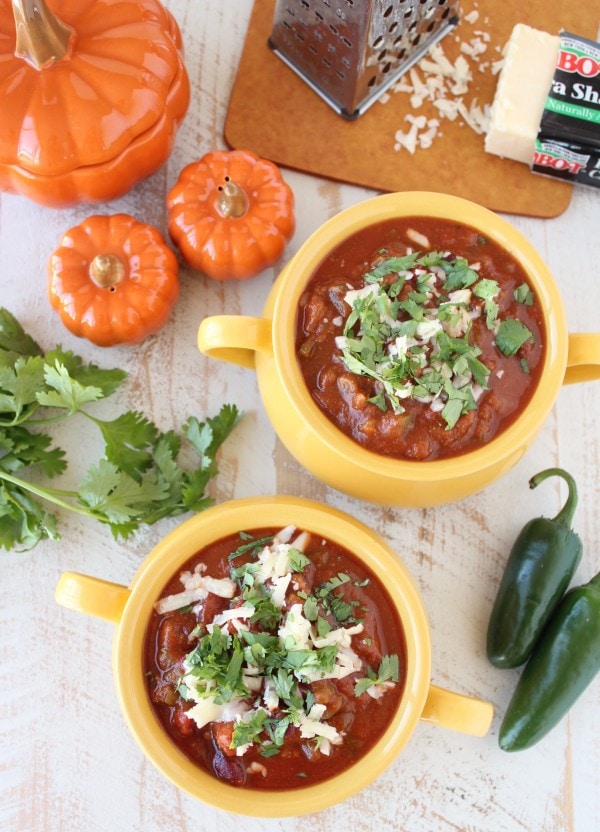 Two yellow bowls filled with Pumpkin Chili garnished with melted cheese and cilantro on a wooden surface surrounded by jalapeños, cilantro, and pumpkins. 