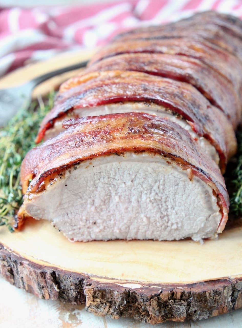 Sliced bacon wrapped pork loin on cutting board with fresh herbs