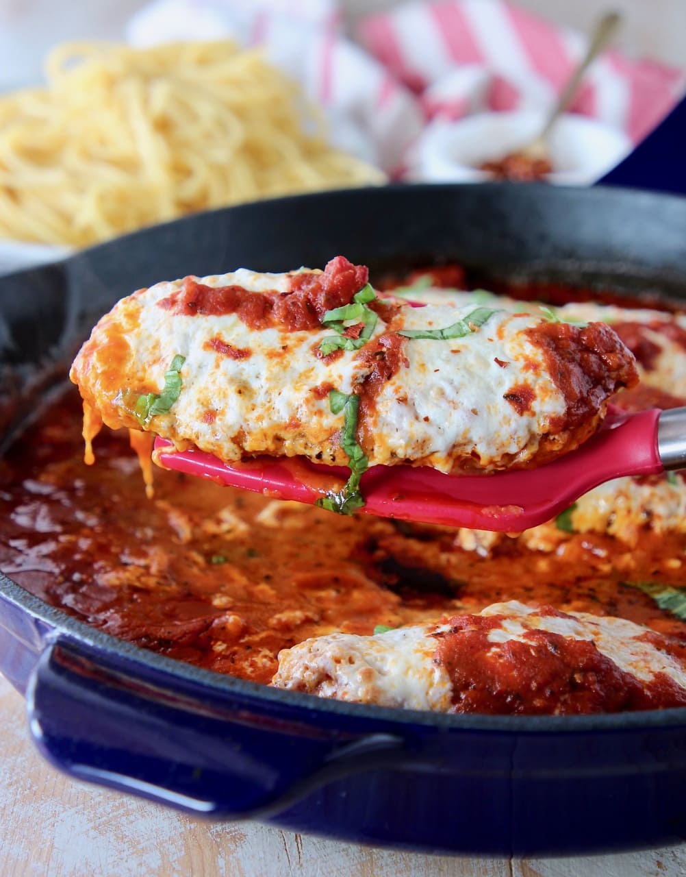 Espátula roja sosteniendo un trozo de pollo a la parmesana saliendo de una sartén azul grande