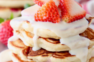 Strawberry pancakes stacked up on white plate with cream cheese syrup and fresh sliced strawberries