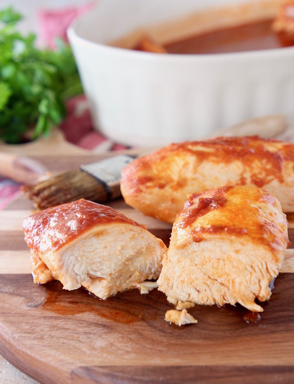 Two pieces of baked chicken on wood cutting board with one piece of chicken sliced in half and pastry brush in the background 
