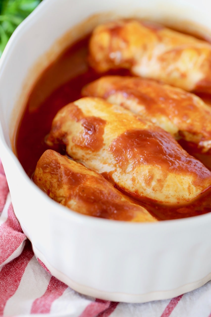 Baked chicken breasts in beige casserole dish, covered in buffalo bbq sauce, sitting on top of red and white striped towel