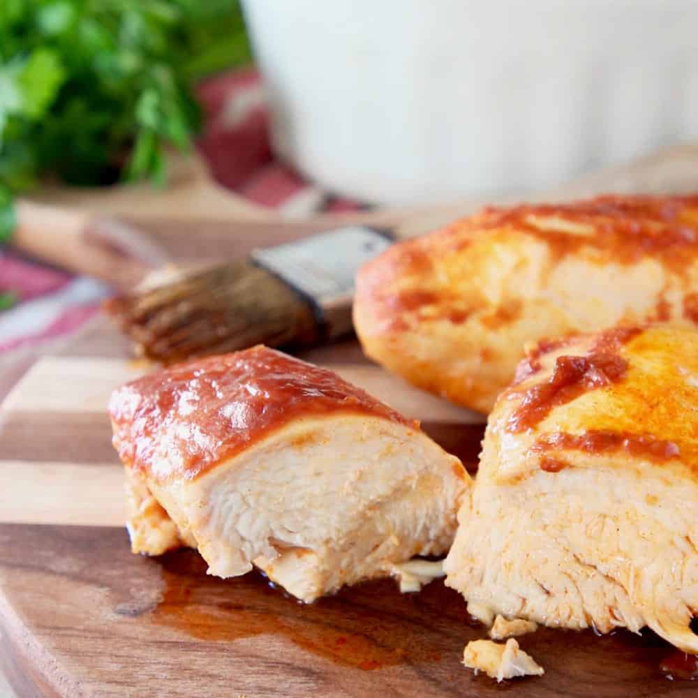 Baked Chicken covered in bbq and buffalo sauce, one piece sliced in half on wood cutting board with pastry brush in background