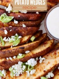 Buffalo-spiced crispy baked potato wedges garnished with green onions and cheese crumble, accompanied by a bowl of creamy dip on the side.