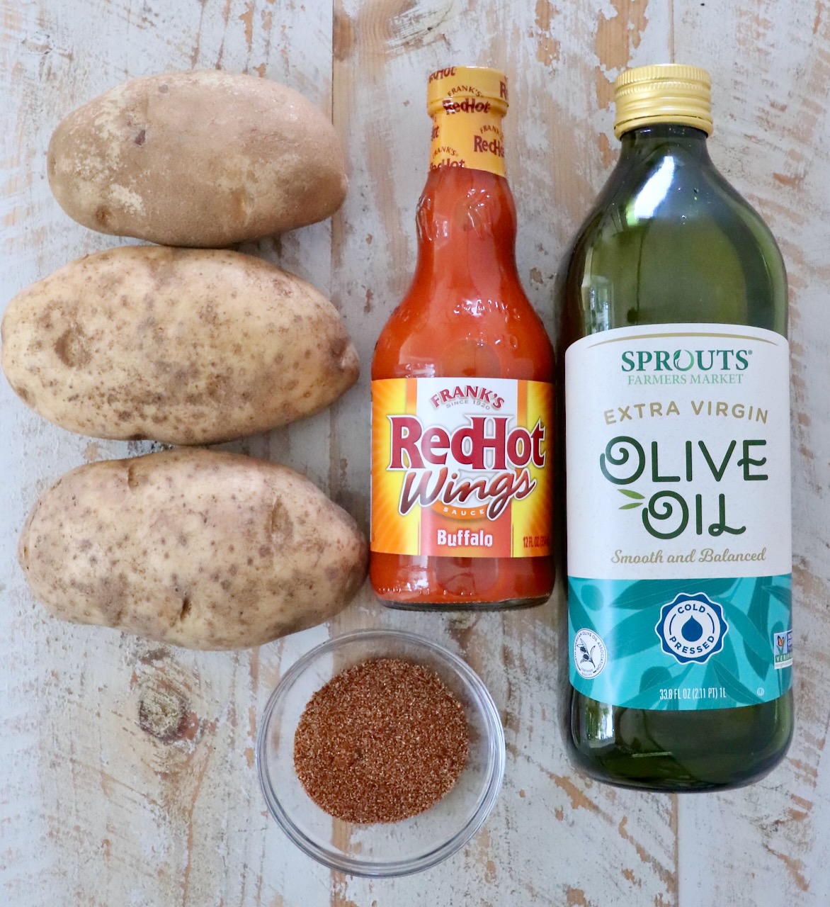 Three potatoes, a bottle of hot sauce, olive oil, and a small bowl of seasoning are arranged on a white wooden surface