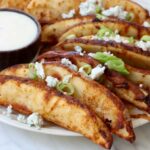 Crispy potato wedges garnished with blue cheese crumbles and green onions on a plate, served with a side of creamy ranch dipping sauce.