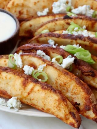 Crispy potato wedges garnished with blue cheese crumbles and green onions on a plate, served with a side of creamy ranch dipping sauce.
