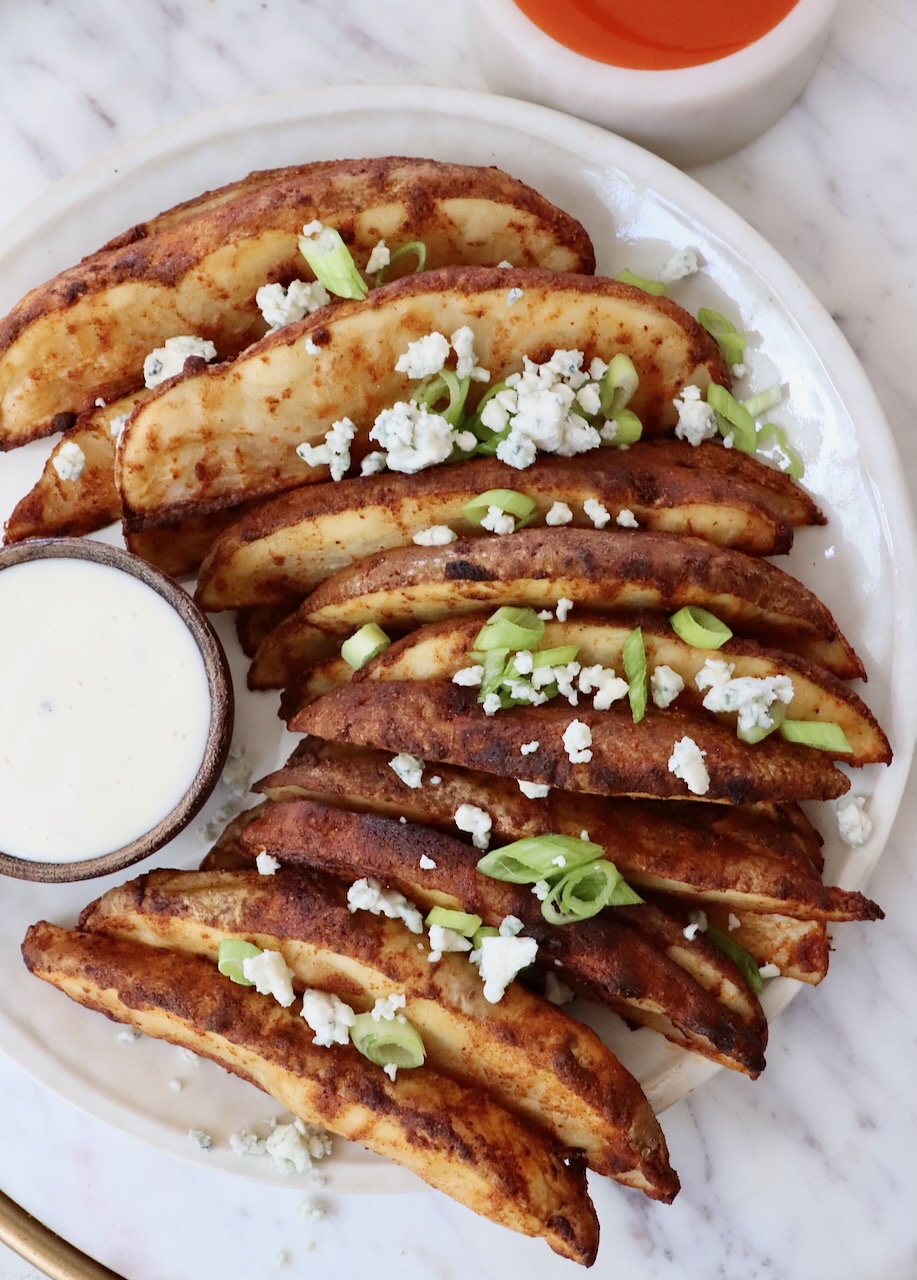 Crispy potato wedges on a plate, topped with green onions and crumbled blue cheese. A bowl of creamy dip sits beside them on the plate.
