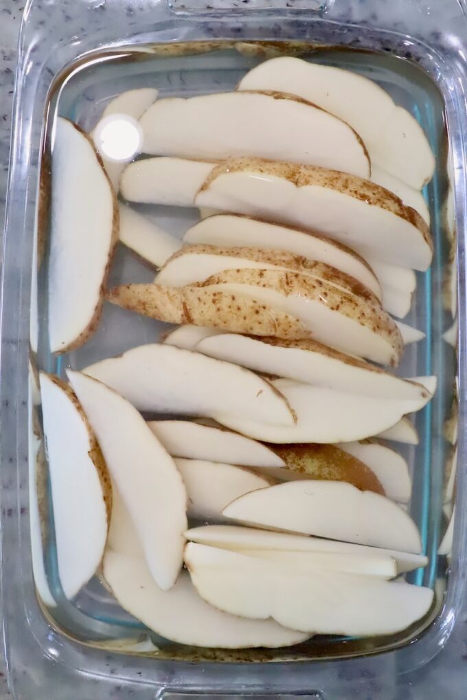 Sliced potatoes soaking in a clear glass dish filled with water on a marble countertop.