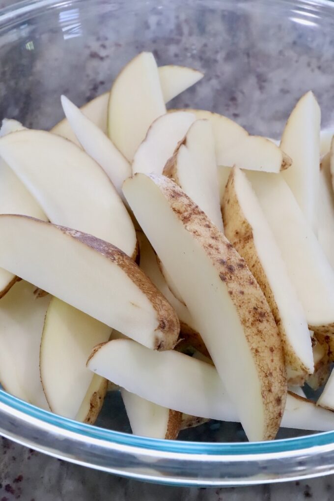 A glass bowl filled with raw potato wedges, showcasing their light brown skins and creamy flesh. The slices are arranged haphazardly.