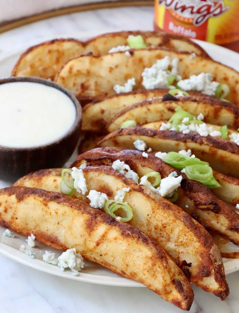 Crispy potato wedges garnished with blue cheese crumbles and green onions on a plate, served with a side of creamy ranch dipping sauce.