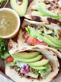 Three ground beef tacos on wood tray with avocado and salsa