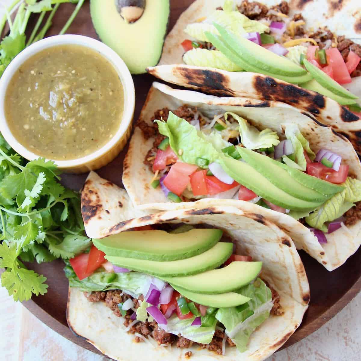 Three ground beef tacos on wood tray with avocado and salsa