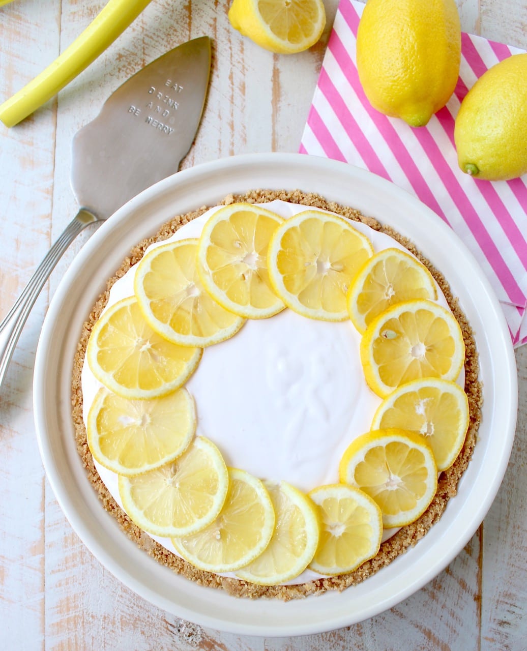 A creamy lemon pie topped with lemon slices sits on a wooden table. Beside it are whole lemons, a striped napkin, and a pie server.