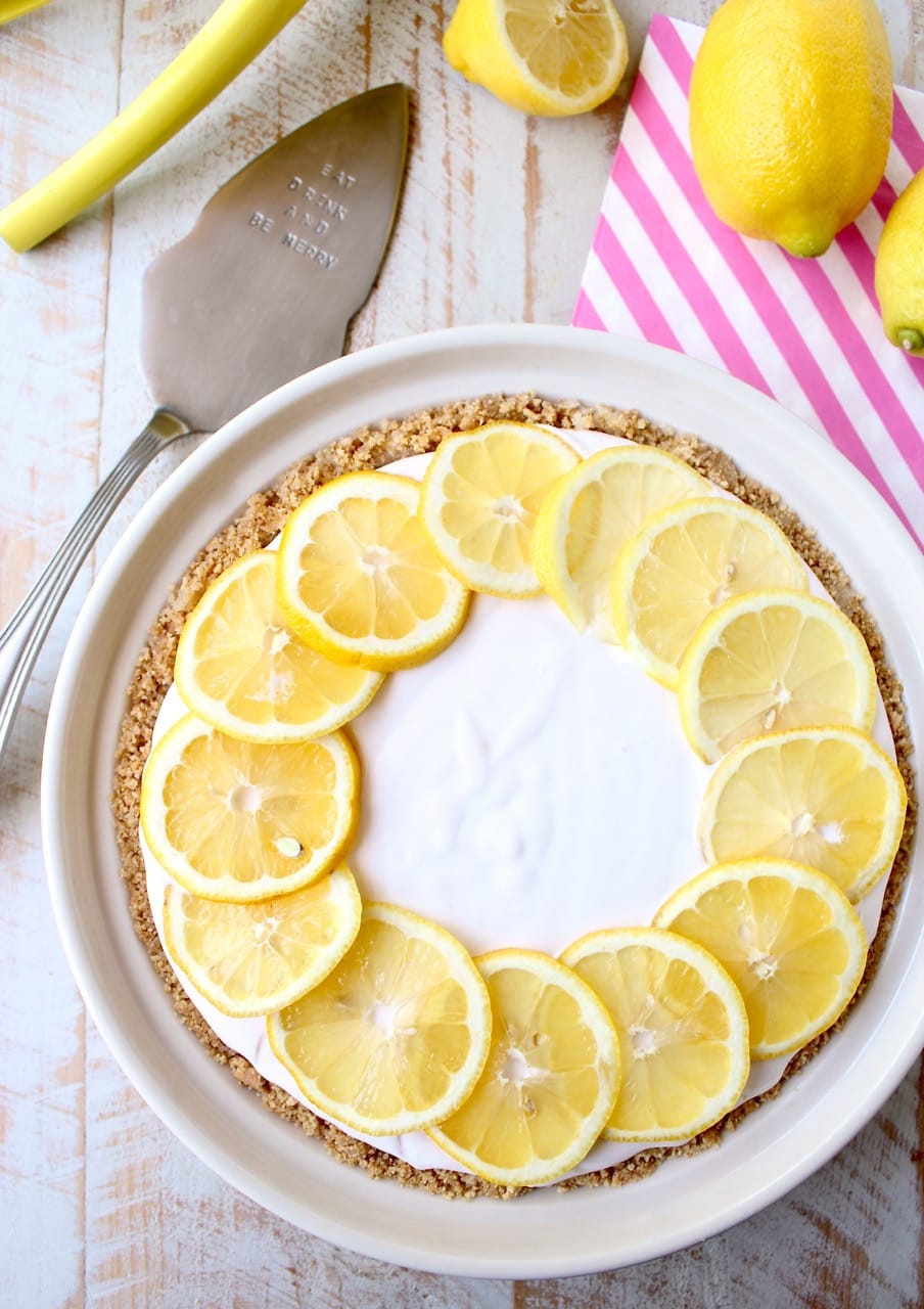 A creamy lemonade pie topped with lemon slices sits on a wooden table. Beside it are whole lemons, a striped napkin, and a pie server.