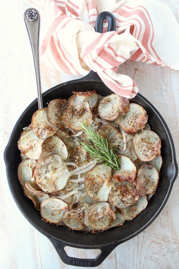 A cast iron skillet on a wooden surface filled with sliced and seasoned potatoes with a fork.