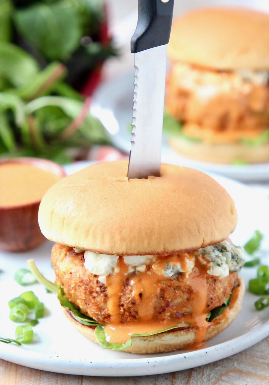 Buffalo chicken burger on plate, topped with blue cheese crumbles with knife in the middle of the burger