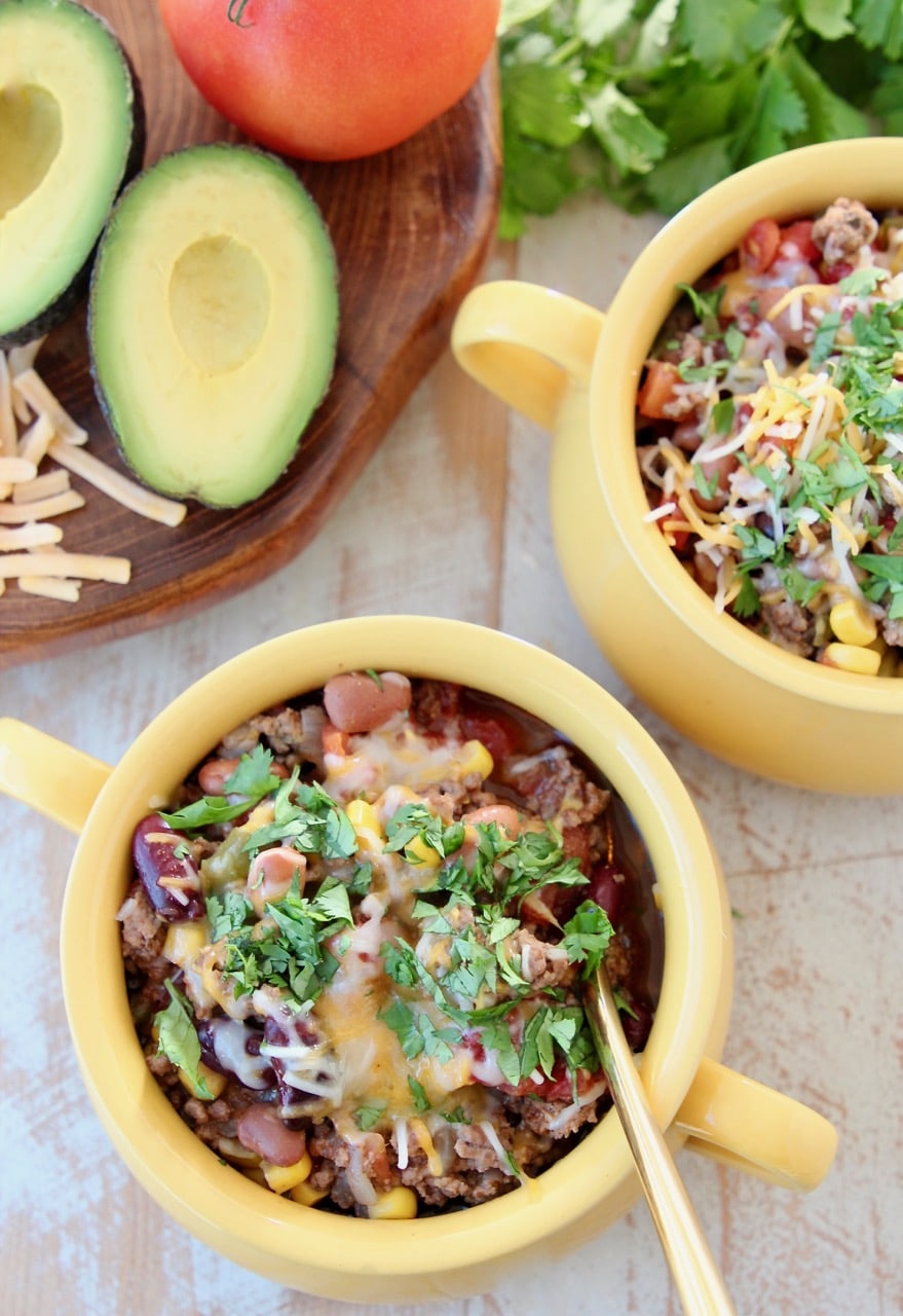 Taco soup in yellow crocks with gold spoon, fresh cilantro, cheddar cheese and fresh avocado