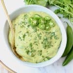 Creamy green salsa in bowl with gold spoon and serrano peppers