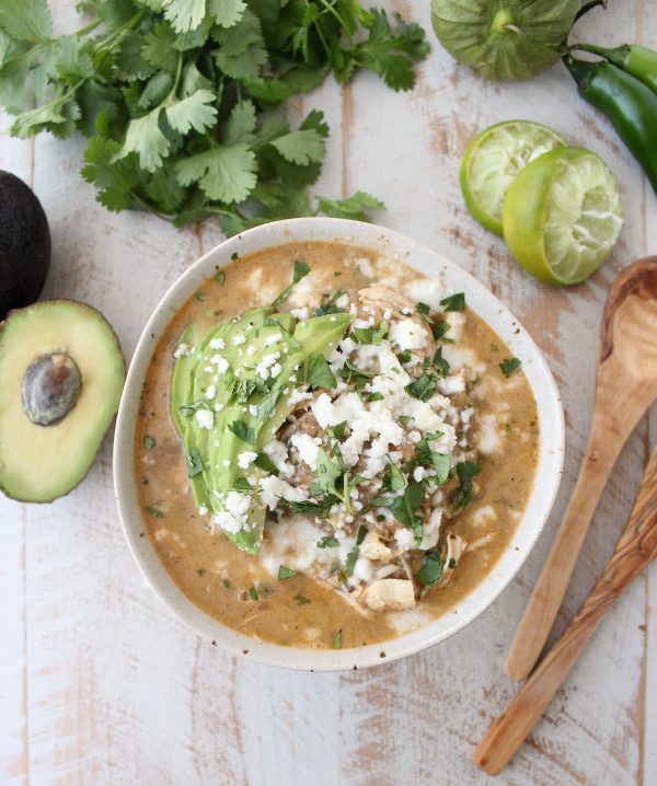 Green Chili Chicken Enchilada Soup topped with avocado, cilantro, and cheese on a wooden surface.