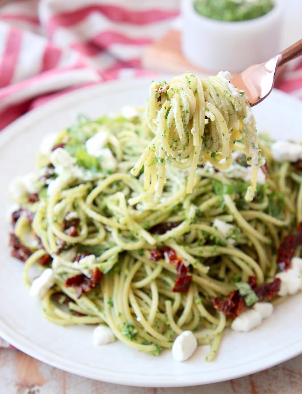 Pesto spaghetti wrapped around a copper fork, held over a plate of pesto spaghetti with crumbled goat cheese and sun dried tomatoes