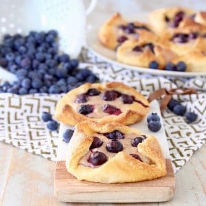 Blueberry Pastries sitting on cutting board on top of gold and white patterned towel with a white colander of blueberries spilling on in the background next to a plate of blueberry pastries
