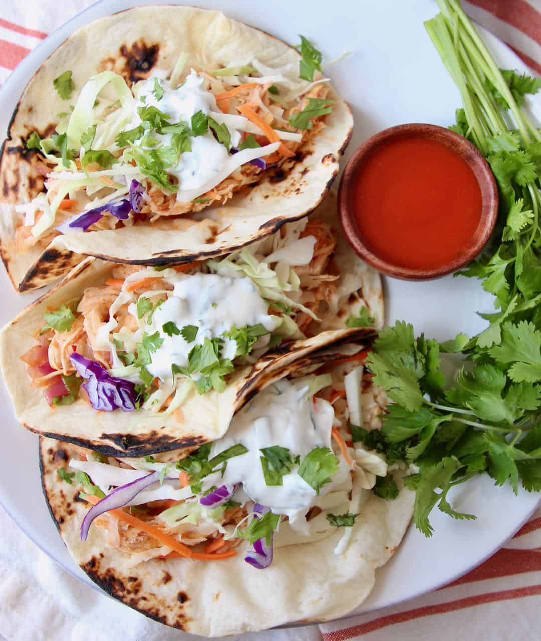 Three buffalo chicken tacos on white plate with side small bowl of buffalo sauce