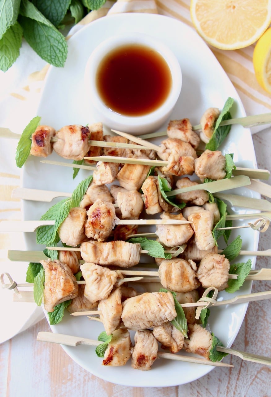 Overhead shot of chicken skewers on white plate with small bowl of bourbon sauce on the side