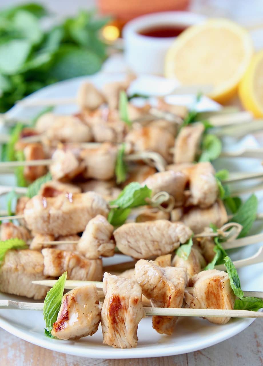 Grilled chicken pieces on small wooden skewers with fresh mint leaves, sitting on a white oval plate