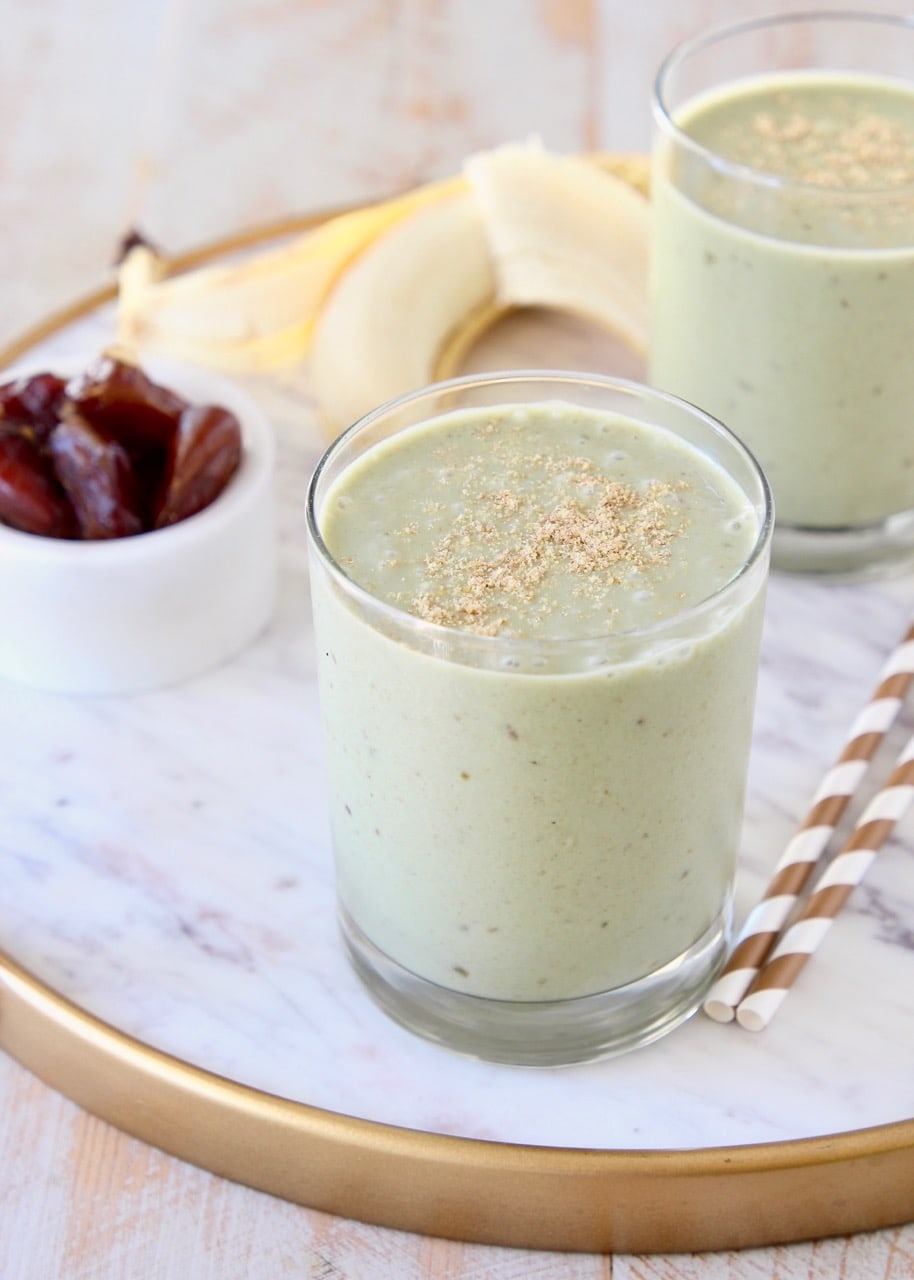 Banana protein shake in two glasses sitting on white marble tray with gold and white striped straws and peeled banana in background