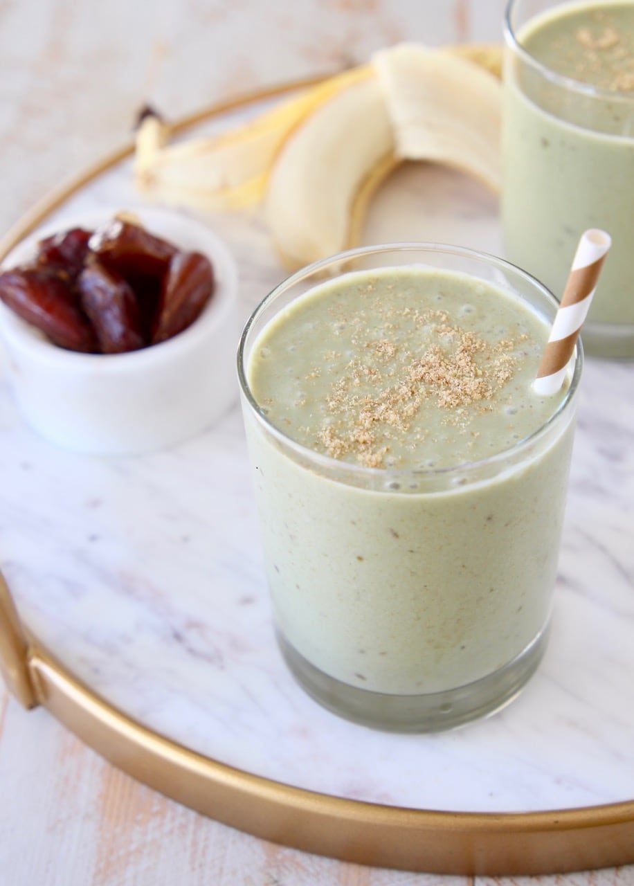 Banana protein shake in glass with gold and white striped straw and ground flaxseed on top with peeled banana in background