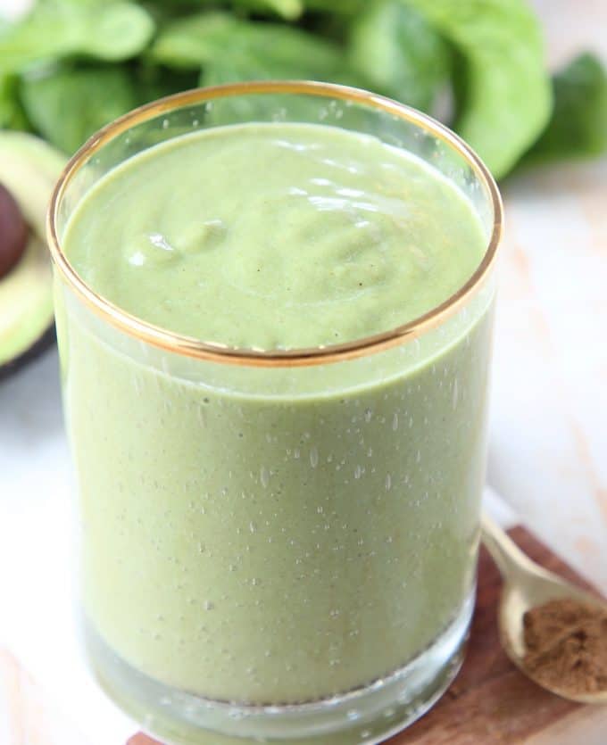 Green smoothie in glass sitting in front of fresh spinach and an avocado