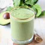 Green smoothie in glass, sitting next to an avocado half and fresh spinach