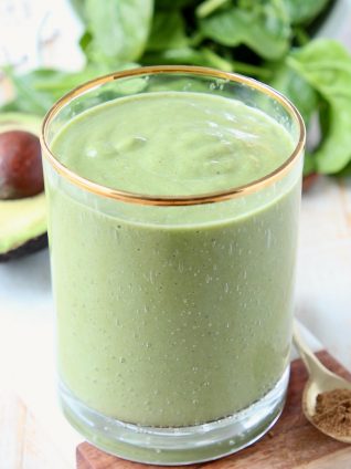 Green smoothie in glass, sitting next to an avocado half and fresh spinach