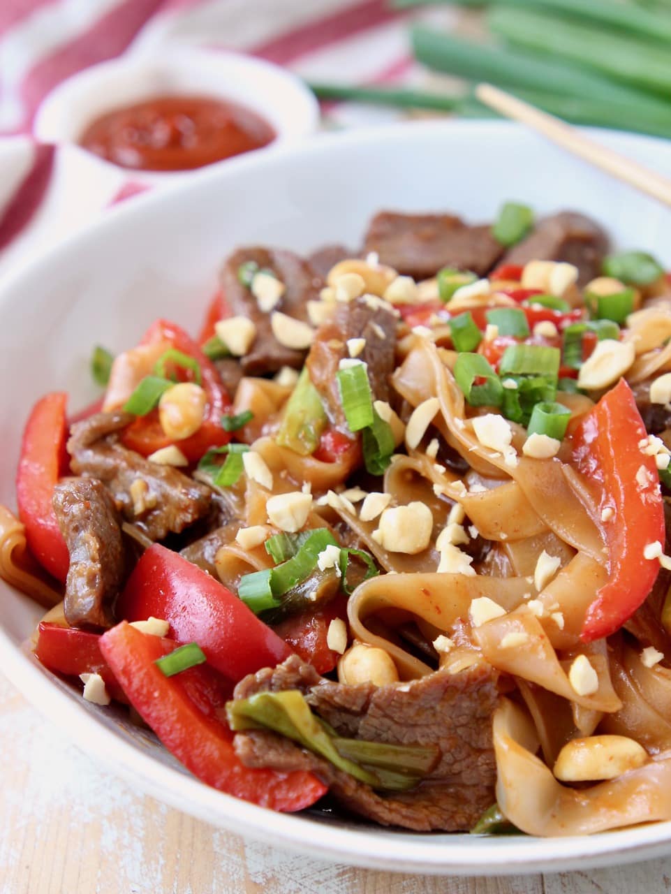 Kung pao beef, rice noodles and red bell peppers in white bowl topped with peanuts and green onions