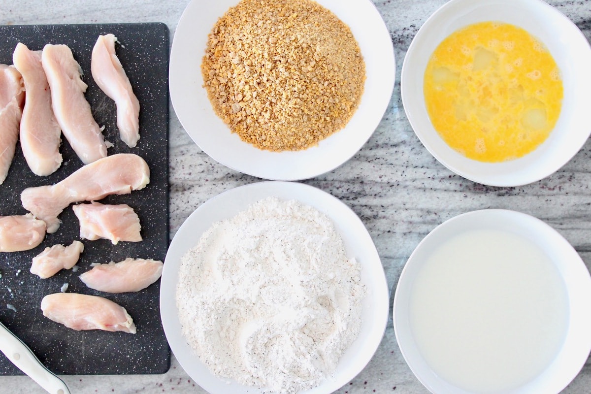 Chicken strip batter station with bowl of eggs, bowl of milk, plate of flour, plate of crushed cereal and raw chicken on cutting board
