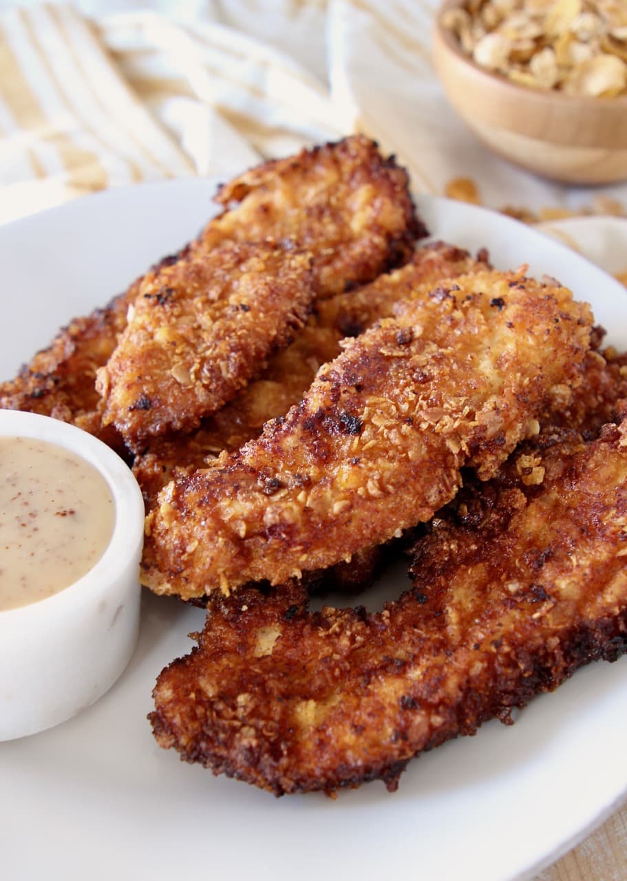 Honey bunches of oats fried chicken strips on plate with a ramekin of honey mustard dressing on yellow and white striped towel