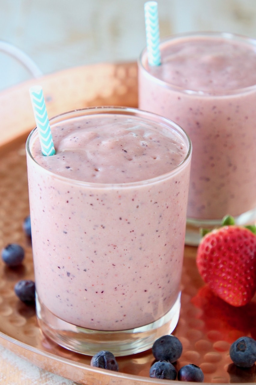 Fruit smoothie in glass with blue and white striped straw on copper tray with fresh blueberries and strawberries