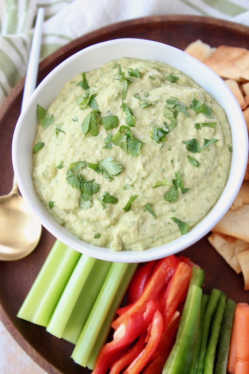 Spinach artichoke hummus in white bowl on wood serving tray with fresh celery, carrots and bell peppers