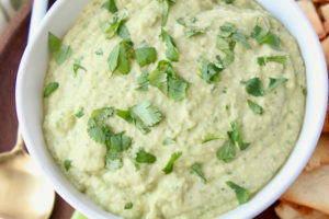 Spinach artichoke hummus in white bowl with gold and white spoon and pita chips on the side