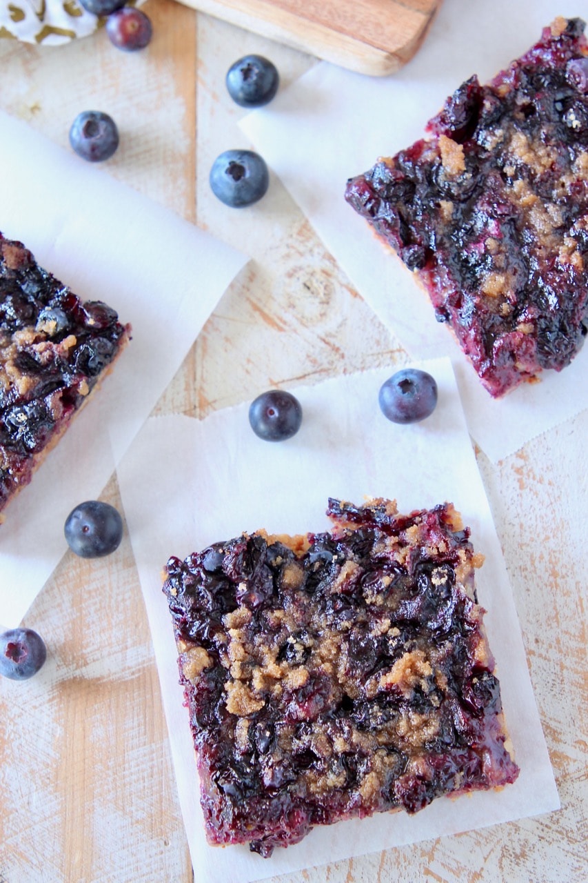 Overhead shot of blueberry oat bars cut into squares, sitting on white parchment paper with fresh blueberries