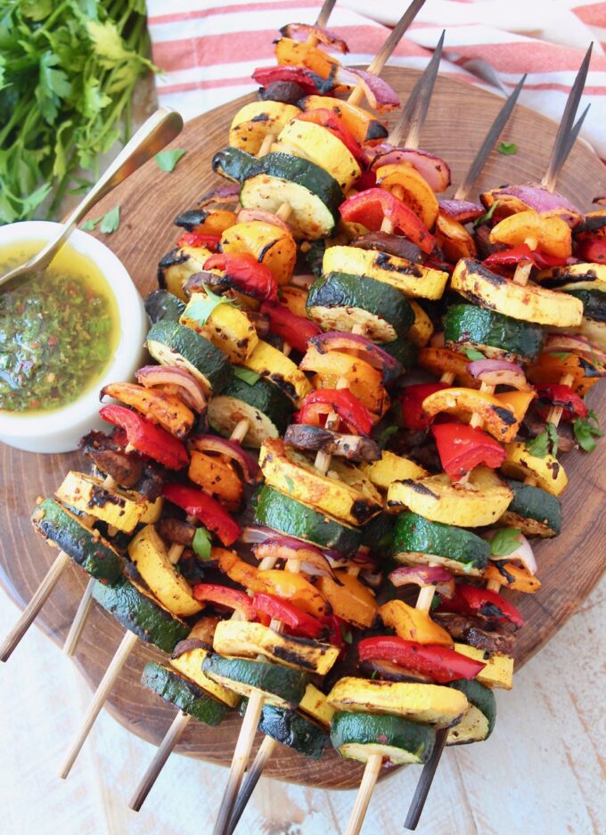 skewers of grilled vegetables on wood serving tray with chimichurri sauce on the side