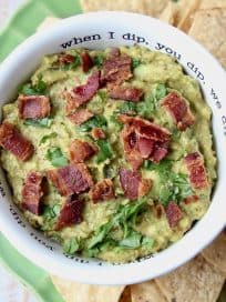 Guacamole in white bowl topped with cooked bacon, with tortilla chips on the side