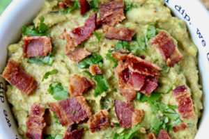 Guacamole in white bowl topped with cooked bacon pieces and fresh cilantro