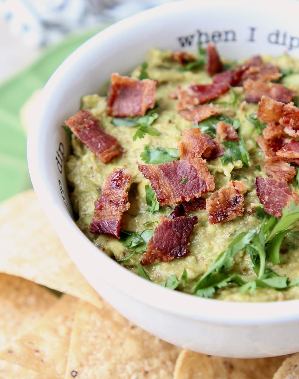 Guacamole in white bowl topped with diced cooked bacon pieces and fresh cilantro