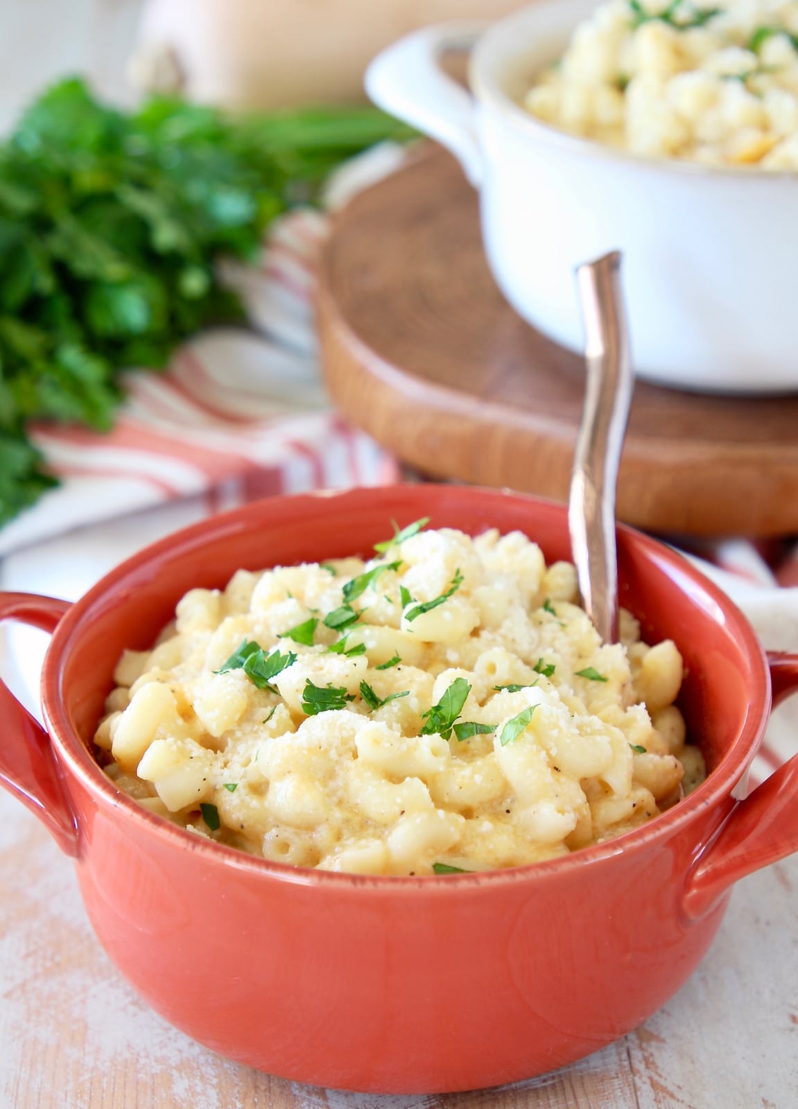 Butternut squash mac and cheese in orange crock with copper spoon, with white crock of mac and cheese in background and fresh parsley