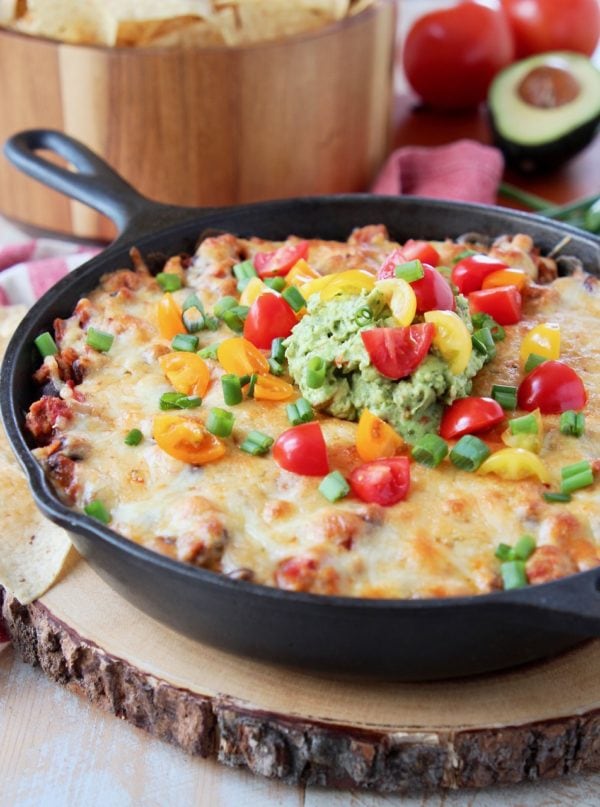 Baked taco dip in a cast iron skillet, sitting on a wood cutting board with a bowl of chips in the background, sitting on a red and white striped towel