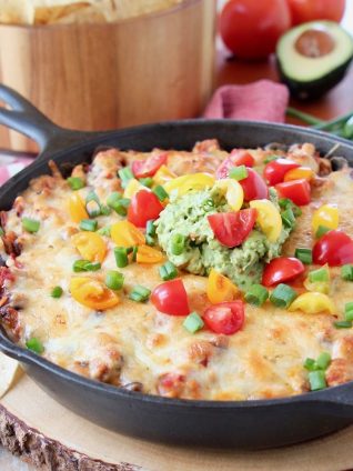 Baked taco dip in a cast iron skillet, sitting on a wood cutting board with a bowl of chips in the background