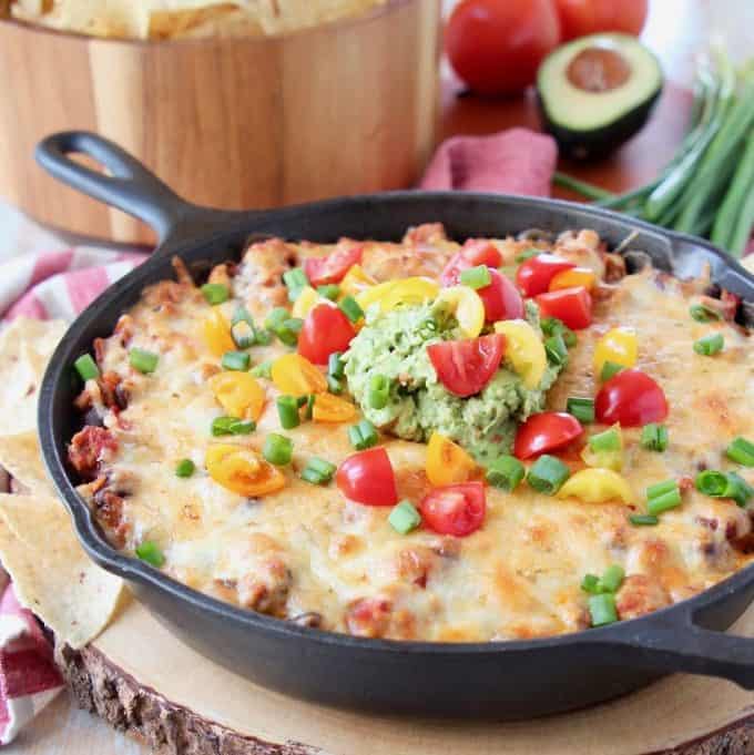 Baked taco dip in a cast iron skillet, sitting on a wood cutting board with a bowl of chips in the background