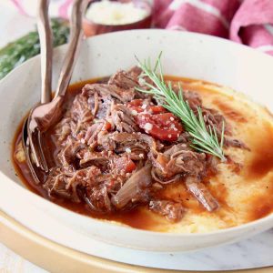Shredded beef ragu tomato sauce in bowl with creamy polenta and rosemary sprig